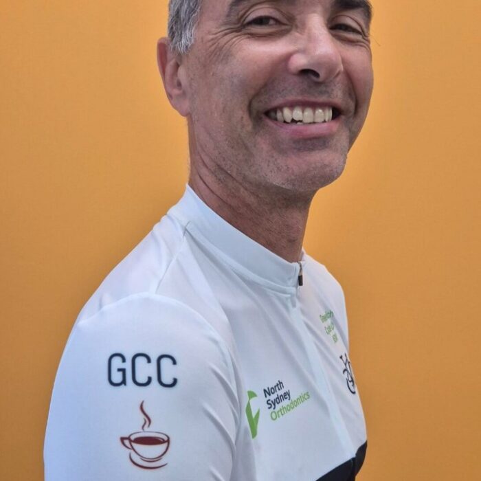 20251029_135917 Smiling man in a white cycling jersey (sleeve shows GCC and a coffee cup icon) photographed in front of an orange background, shown in profile.