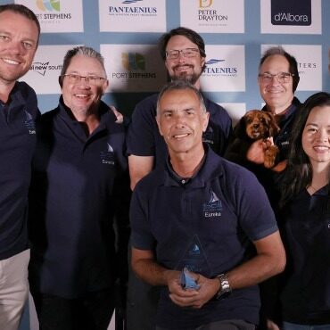 IMG-20260419-WA0003 Group of seven people posing together on a blue sponsor backdrop; a man in front holds a trophy, a small dog sits with another person nearby.