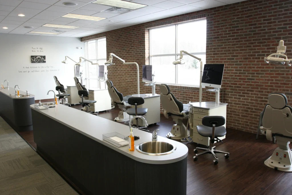 open plan orthodontic clinic - North Sydney Orthodontics A modern dental clinic with multiple treatment chairs, overhead lights, and computer monitors along a brick wall and large windows.